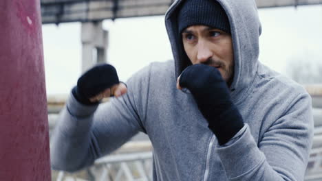 Close-up-view-of-caucasian-man-in-grey-hoodie-hitting-a-punching-bag-outdoors-an-abandoned-factory-on-a-cloudy-morning