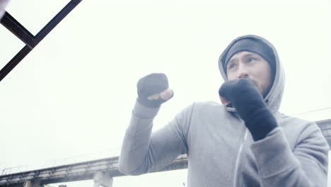 Bottom-view-of-caucasian-man-in-grey-hoodie-hitting-a-punching-bag-outdoors-an-abandoned-factory-on-a-cloudy-morning