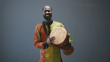 Porträt-Eines-Jungen-Fröhlichen-Afroamerikanischen-Mannes-In-Traditioneller-Kleidung,-Der-Einen-Schlagzeuger-Spielt-Und-In-Die-Kamera-Lächelt