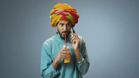 Young-cheerful-Indian-man-in-traditional-clothes-and-turban-talking-on-the-phone-and-drinking-orange-juice