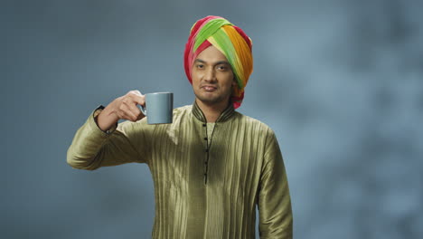 Portrait-of-young-happy-indian-man-in-turban-and-traditional-clothes-drinking-tea-or-coffee-and-smiling-at-camera