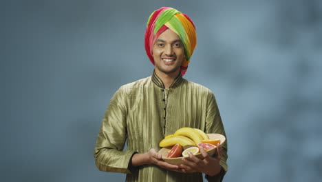 Young-happy-indian-man-in-traditional-clothes-and-turban-smiling-cheerfully-at-camera-and-holding-plate-with-fruits