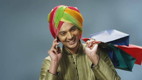Close-up-view-of-cheerful-Indian-man-in-traditional-clothes-and-turban-talking-on-the-phone-and-holding-shopping-bags
