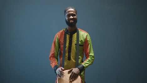 Porträt-Eines-Jungen-Fröhlichen-Afroamerikanischen-Mannes-In-Traditioneller-Kleidung,-Der-Einen-Schlagzeuger-Spielt-Und-In-Die-Kamera-Lächelt