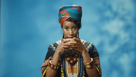 African-American-young-beautiful-woman-in-traditional-clothes-and-turban-drinking-hot-tea-and-looking-at-camera