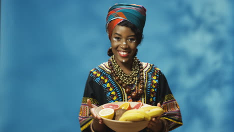Joven-Afroamericana-Alegre-Con-Turbante-Y-Maquillaje-Sonriendo-Alegremente-A-La-Cámara-Mientras-Baila-Y-Sostiene-Un-Plato-Con-Frutas