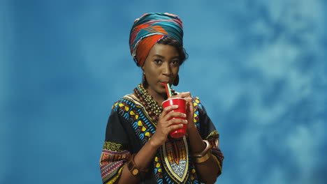 Afroamerikanische-Junge-Frau-In-Turban-Und-Traditioneller-Kleidung,-Die-Saft-Oder-Limonade-Trinkt-Und-In-Die-Kamera-Lächelt