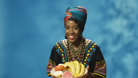 Joven-Afroamericana-Alegre-Con-Turbante-Y-Ropa-Tradicional-Sonriendo-Alegremente-A-La-Cámara-Mientras-Baila-Y-Sostiene-Un-Plato-Con-Frutas