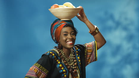 African-American-young-cheerful-woman-in-turban-and-traditional-clothes-smiling-cheerfully-at-camera-while-holding-a-plate-with-fruits-on-her-head