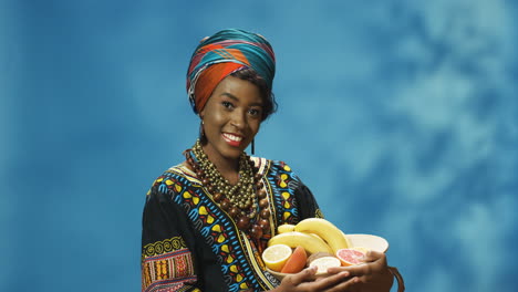 Joven-Afroamericana-Alegre-Con-Turbante-Y-Ropa-Tradicional-Sonriendo-Alegremente-A-La-Cámara-Mientras-Sostiene-Un-Plato-Con-Frutas