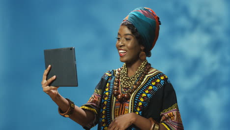 African-American-young-cheerful-woman-in-turban-and-traditional-clothes-making-a-video-call-using-a-tablet
