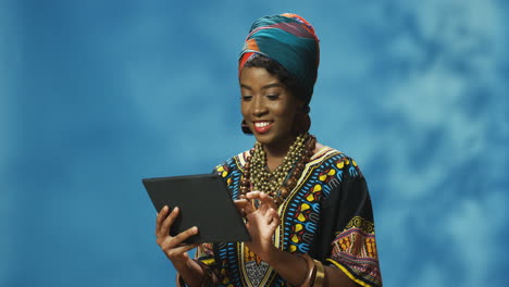 African-American-young-cheerful-woman-in-turban-and-traditional-clothes-holding-tablet-and-tapping-or-scrolling-on-the-screen