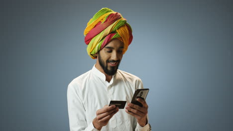 Junger-Fröhlicher-Inder-In-Traditioneller-Kleidung-Und-Turban,-Der-Kreditkarte-Und-Smartphone-In-Den-Händen-Hält-Und-Online-Einkauft