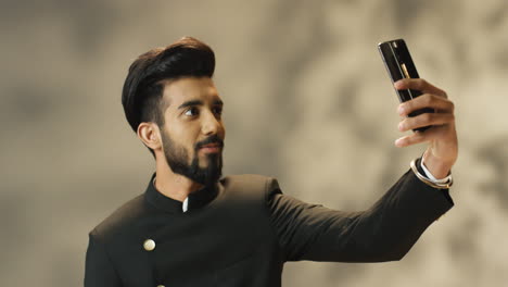 Young-cheerful-Indian-man-in-traditional-clothes-making-a-selfie-with-smartphone