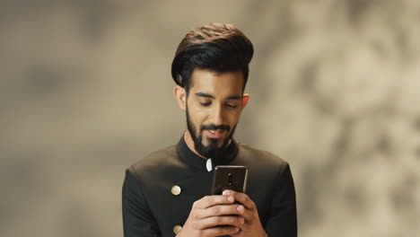 Junger,-Fröhlicher-Indischer-Mann-Mit-Bart-Und-In-Schwarzer-Jacke,-Der-Lächelt,-Während-Er-Auf-Dem-Smartphone-SMS-Schreibt
