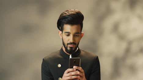 Young-cheerful-Indian-man-with-beard-and-in-black-jacket-texting-on-smartphone