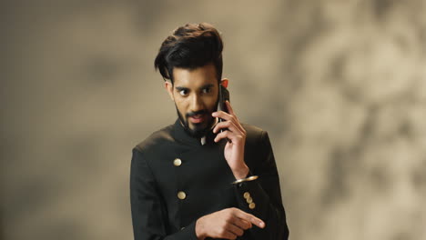 Young-cheerful-Indian-man-with-beard-and-in-black-jacket-talking-on-smartphone