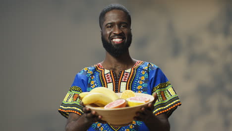 Portrait-of-young-cheerful-African-American-man-in-traditional-clothes-holding-and-showing-a-plate-with-tropical-fruit-while-looking-at-camera