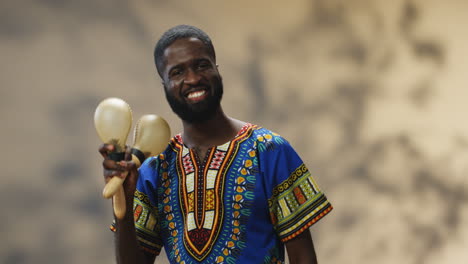 Junger-Fröhlicher-Afroamerikanischer-Mann-In-Traditioneller-Kleidung,-Der-Maracas-Spielt-Und-In-Die-Kamera-Lächelt