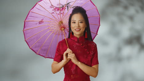 Porträtaufnahme-Einer-Jungen,-Fröhlichen-Asiatin-In-Roter-Traditioneller-Kleidung,-Die-Einen-Sonnenschirm-Hält-Und-In-Die-Kamera-Lächelt