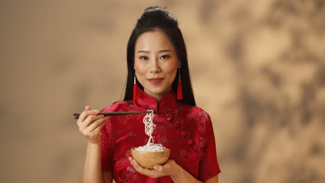 Portrait-shot-of-Asian-young-cheerful-woman-in-red-traditional-clothes-eating-noodles-and-smiling-at-camera