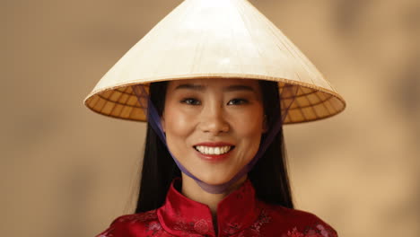 Close-up-view-of-young-Asian-happy-woman-in-red-clothes-and-conus-hat-smiling-joyfully-at-camera