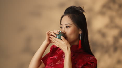 Close-up-view-of-young-Asian-woman-in-red-traditional-clothes-smiling-cheerfully-at-camera-and-drinking-tea-from-a-cup