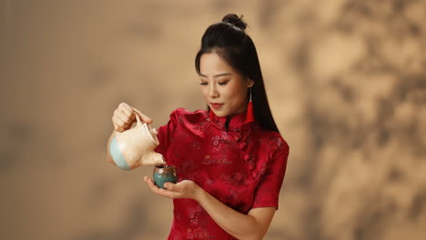 Young-Asian-woman-in-red-traditional-clothes-pouring-hot-tea-from-ceramic-kettle-to-cup-and-smiling-at-camera