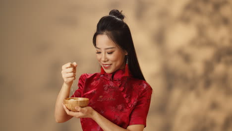 Cheerful-young-Asian-woman-in-red-traditional-costume-holding-plate-and-pouring-rice-from-hand