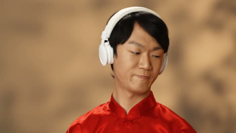 Close-up-view-of-cheerful-young-Asian-man-in-red-traditional-costume-wearing-headphones-and-listening-to-music