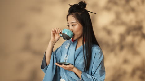 Asian-young-woman-in-blue-kimono-drinking-tea-from-a-ceramic-cup-and-smiling-at-camera