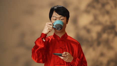Close-up-view-of-cheerful-young-Asian-man-in-red-traditional-costume-drinking-tea-from-a-ceramic-cup-and-looking-at-camera