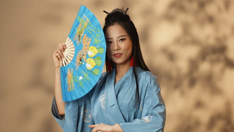 Junge-Asiatische-Frau-Im-Blauen-Kimono-Winkt-Mit-Einem-Fan-Und-Versteckt-Ihr-Gesicht,-Während-Sie-Lächelt-Und-Flirtet