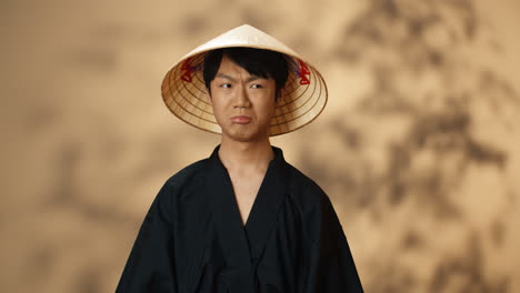 Junger-Asiatischer-Mann-In-Schwarzer-Traditioneller-Tracht-Und-Konushut,-Der-Mit-Wütendem-Gesicht-In-Die-Kamera-Blickt