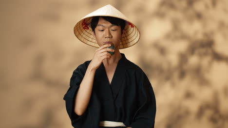 Young-Asian-man-in-black-traditional-costume-and-conus-hat-drinking-tea-and-smiling-at-camera