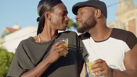 Portrait-of-carefree-gays-kissing-and-drinking-juice-outside