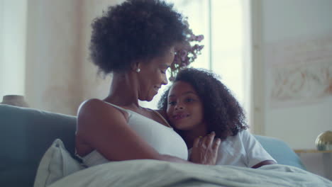 Medium-shot-of-happy-African-American-mum-and-daughter-lying-in-bed-and-talking-together