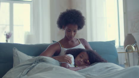 Dolly-shot-of-a-caring-African-American-mum-stroking-her-daughter's-hair-while-girl-falling-asleep