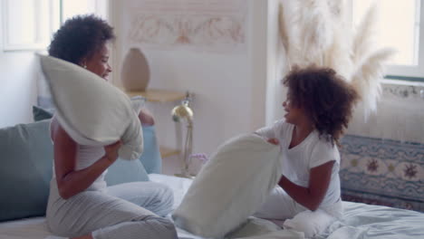 Happy-African-American-mum-and-daughter-having-pillow-fight-in-bed