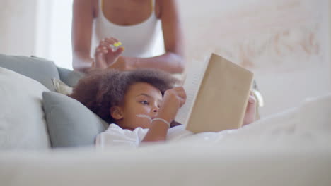Little-African-American-girl-spending-time-reading-in-bed-when-her-mum-coming-up,-smiling-and-kissing-her