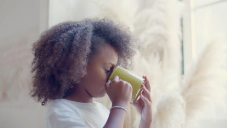 Cute-Black-girl-standing-by-the-window,-drinking-tea,-then-turning-to-camera-and-smiling
