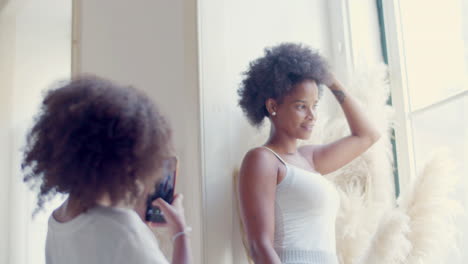 Black-woman-posing-by-window-while-her-little-daughter-taking-photo-and-then-showing-it-to-mum