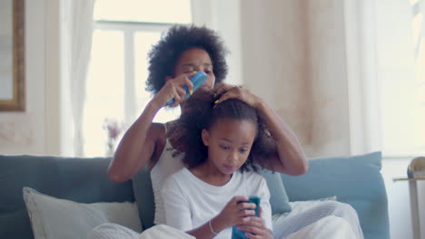 Cute-Black-daughter-playing-mobile-game-on-her-smartphone-sitting-on-bed-while-mum-combing-her-hair