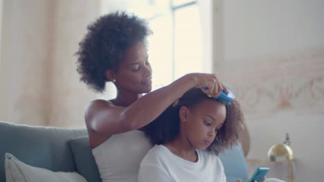 African-American-mum-combing-her-daughter's-hair-sitting-on-couch-while-little-girl-using-mobile-phone