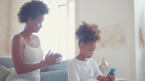 African-American-mum-combing-her-daughter's-hair-while-little-girl-using-listening-music-on-phone-and-dancing