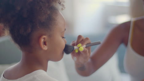 Close-up-of-a-little-African-American-girl-having-her-mum-doing-make-up-on-her,-applying-powder-on-her-cheeks-with-makeup-brush