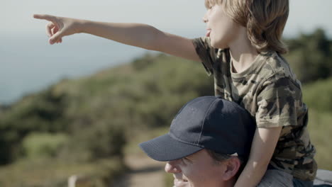 Close-up-of-father-carrying-son-on-shoulders-on-hiking-adventure-while-boy-pointing-at-something-in-distance