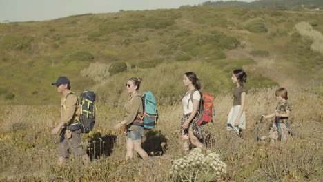 Side-view-of-a-happy-family-with-backpacks-walking-along-hiking-trail-in-row