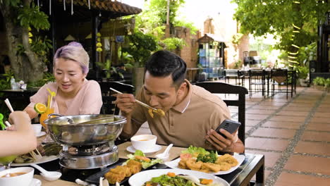 Young-chinese-man-looking-at-this-phone-and-eating-shrimps-while-his-family-is-eating-and-talking