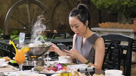 Young-chinese-woman-eating-from-hot-pot-and-using-her-smartphone-at-the-restaurant
